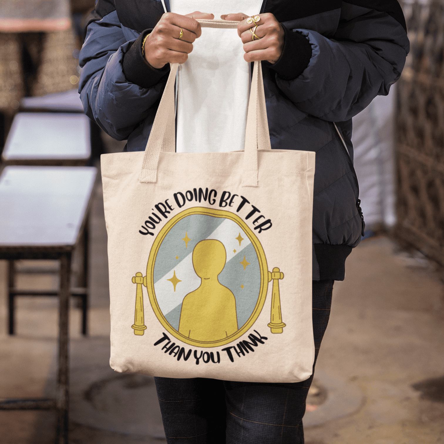 Bolsa de lona ecológica beige con diseño motivacional "You're doing better than you think" y espejo dorado, llevada por persona con chaqueta azul marino